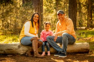 parents-and-daughter-sitting-on-log-in-sunny-forest-smiling